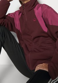 Maroon zip-up jacket with pink nylon accents and high collar. Features a small embroidered logo and side pockets. Smooth texture.