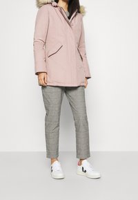 Pink parka with a faux fur hood, featuring front pockets, a zip and button closure, worn over plaid trousers and white sneakers.