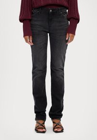 Jeans de mezclilla negros con un corte recto, diseño de cinco bolsillos y sutiles acentos de costura en la parte delantera. Llevados con un suéter burdeos y sandalias.