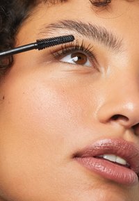 Spazzola di mascara nero che applica il prodotto su lunghe ciglia scure sopra un occhio marrone a forma di mandorla, con pelle liscia e luminosa e un colore delle labbra tenue.