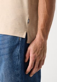 Beige kortermet T-skjorte med sidesplitt, subtil tekstur, stoffetikett sydd på venstre side, brukt med blå denim jeans.