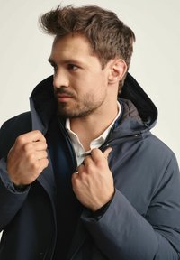 Granatowa kurtka puchowa z kapturem, z zamek błyskawicznym z przodu, teksturowanym podkładem i dopasowanym krojem. Ręce trzymające kołnierz.