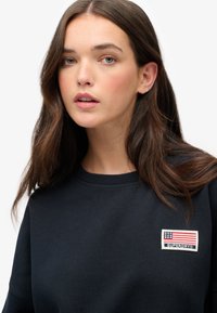 Black sweatshirt with ribbed neckline, featuring an embroidered patch of the American flag and "SUPERDRY" label on the chest.