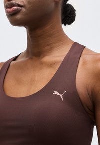 Κοντινό πλάνο ενός ατόμου που φοράει καφέ αθλητικό τοπ με λευκό λογότυπο Puma στο στήθος, τα μαλλιά δεμένα κότσο.