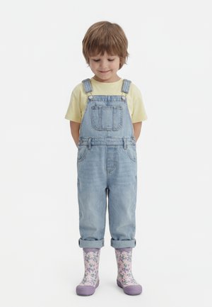 Jeune enfant portant un t-shirt jaune, une salopette en denim bleu clair et des bottes de pluie violettes à motifs floraux, debout, les mains derrière le dos, regardant vers le bas.