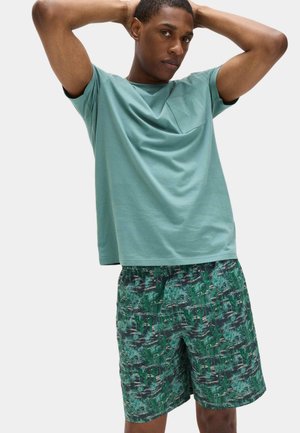 Junger Mann mit erhobenen Armen, der ein hellgrünes T-Shirt und dunkelgrüne Shorts mit tropischem Muster vor einem einfarbigen Hintergrund trägt.