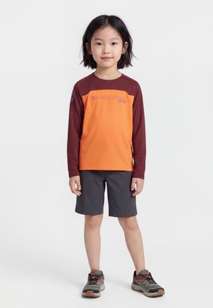 Kind mit kurzen schwarzen Haaren, trägt ein orangefarbenes und kastanienbraunes langärmeliges Shirt, dunkle Shorts und graue Wanderschuhe, steht vor einem weißen Hintergrund.