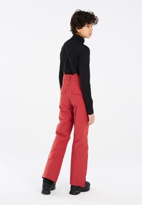 Pantaloni da neve impermeabili rossi con spalline nere regolabili, tasche cargo e gambe larghe, abbinati a un top nero a maniche lunghe e stivali neri.