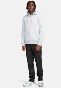 Felpa grigia con zip e finitura texture, dotata di tasca frontale. Abbinata a jeans scuri e sneakers bianche con dettagli blu.
