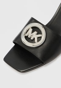 Czarna skórzana sandałka typu slide z szerokim paskiem ozdobionym okrągłym srebrnym logo, kwadratowym noskiem i gładką fakturą.