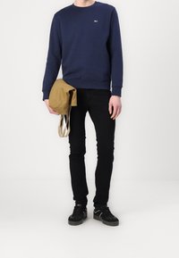 Sudadera azul marino con cuello redondo, vaqueros negros, llevando un bolso beige y calzando zapatillas negras con detalles blancos.