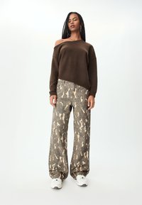 Maglione marrone lavorato a maglia con spalle scoperte abbinato a jeans a gamba larga con motivo mimetico in tonalità beige e verde, completato da sneakers bianche.