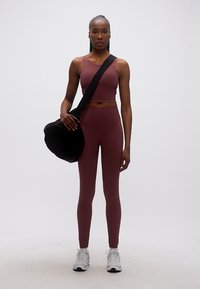 Γυναίκα που στέκεται φορώντας μαρόν αθλητικό κολάν και crop top, κρατώντας μαύρη τσάντα duffel με λευκά αθλητικά παπούτσια σε λευκό φόντο.