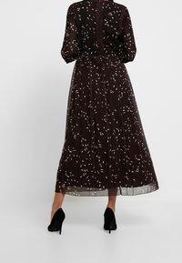 Abito marrone con sovrapposizione trasparente, caratterizzato da un motivo floreale bianco. Maniche lunghe, gonna plissettata e dettaglio con bottoni sul retro.