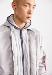 Veste bomber gris clair avec une finition brillante, dotée d'une capuche grise amovible, de fermetures éclair doubles et d'une étiquette en tissu rouge sur la manche.