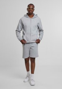 Sweatshirt à capuche gris avec une fermeture éclair sur le devant et une poche kangourou, associé à un short gris assorti. Présenté avec des baskets blanches.