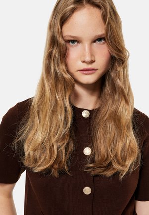 Jeune femme aux longs cheveux blonds ondulés, portant un haut marron foncé avec trois boutons dorés, regardant directement la caméra.