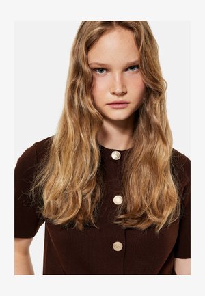 Jonge vrouw met lang golvend blond haar, dragend een donkerbruine top met drie gouden knopen, kijkt recht in de camera.