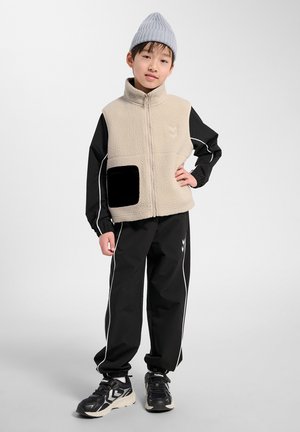 Dziecko w szarej czapce, beżowej kamizelce polarowej, czarnym dresie z białymi lamówkami i czarnych tenisówkach, stojące z jedną ręką na biodrze.