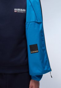 Marineblå sweatshirt med en blå, lynlåset ærme, der har en sort lap og logo. Blød tekstur, ribbet nederkant og ærmer.