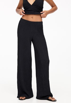 Kobieta nosząca teksturowane czarne spodnie o szerokich nogawkach i dopasowany bezrękawnik crop top, lekko podciąga górę.