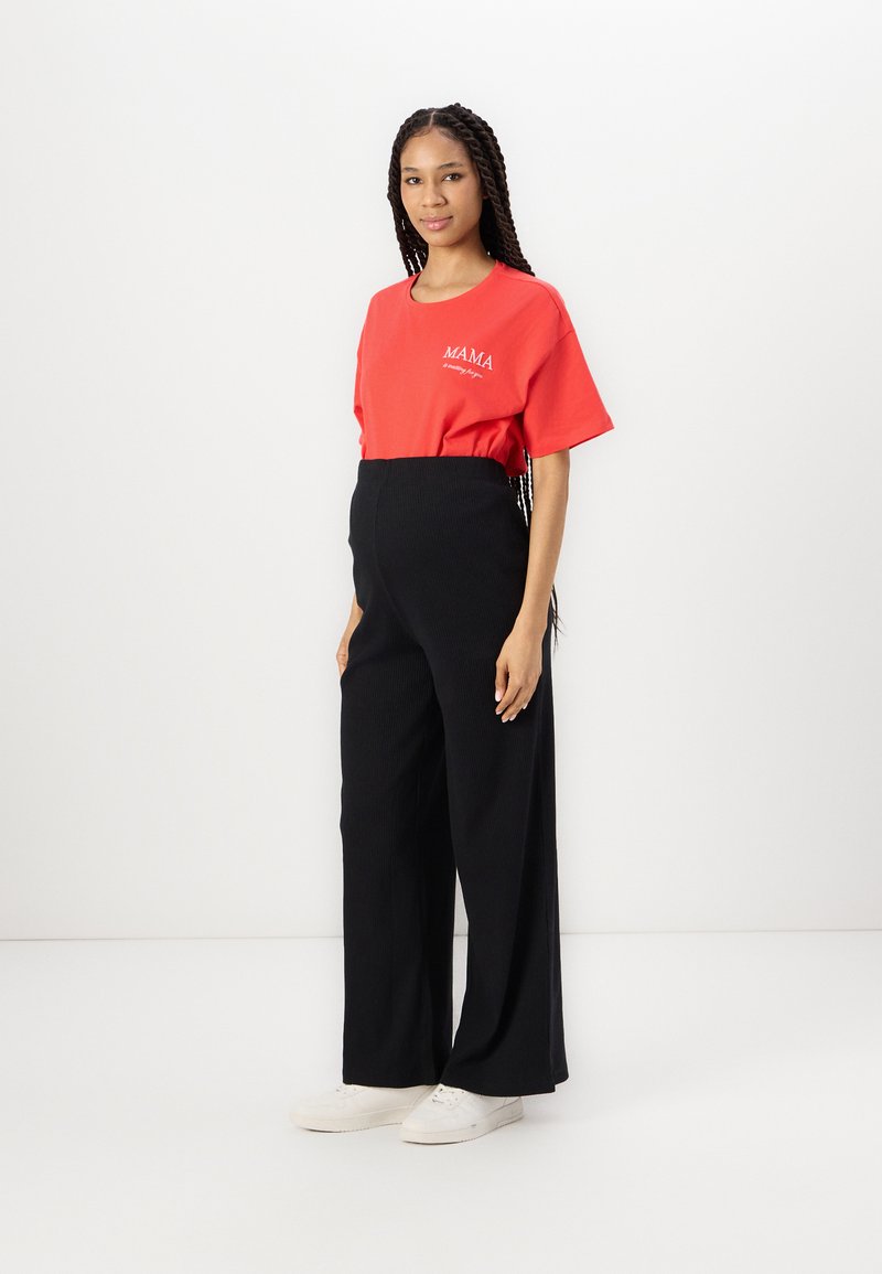 T-shirt rouge à manches courtes avec le texte "MAMA", associé à un pantalon noir à jambes larges. Le mannequin porte des baskets blanches. Fond minimaliste et épuré.