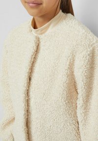 Kremowa, teksturowana kurtka wykonana z puszystego materiału, z okrągłym dekoltem i otwartym przodem. Miękka i przytulna w wyglądzie.