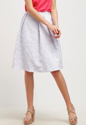Femme portant une jupe A-line blanche à fleurs, un top sans manches rose et des sandales à talons beiges, se tenant devant un fond uni.