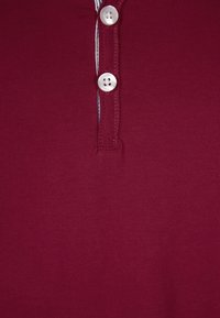 Bordeaux polo tričko s tříknoflíkovým léga a bílými knoflíky, s jedním kontrastním pruhem podél okraje límce. Hladká textura.