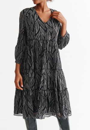 Frau trägt ein schwarzes Midikleid mit weißen abstrakten geschwungenen Blattmustern und dreiviertellangen, gepufften Ärmeln, posiert mit einer Hand in der Nähe des Kragens.