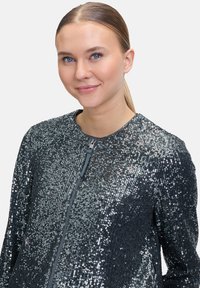 Glitzernde schwarze Jacke mit Reißverschluss vorne, bedeckt mit Pailletten. Sie hat einen runden Ausschnitt und lange Ärmel.