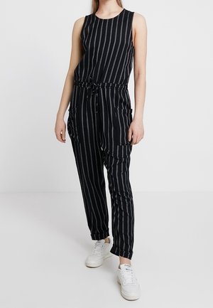 Femme portant une combinaison noire sans manches avec des rayures verticales blanches et des baskets blanches, debout devant un fond blanc uni.