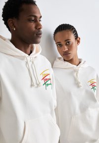 Sudaderas blancas con cordones, que presentan un diseño de logotipo colorido en rojo, amarillo y verde. Textura suave, bolsillos tipo canguro y corte relajado.