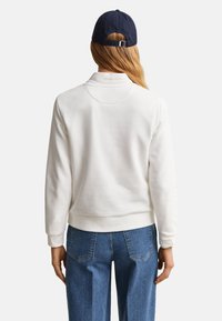 Άτομο με μακριά μαλλιά που φοράει μπλε σκουφάκι baseball, λευκό φούτερ με μακριά μανίκια και μπλε τζιν, στέκεται γυρισμένο με την πλάτη προς τα εμπρός.