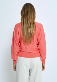 Korallsfärgad tröja med lösa, oversized ärmar och ribbast veck, ihop med ljust färgade byxor, sedd bakifrån med långt hår synligt.