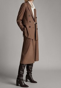 Brąsrowa, w kratę, dopasowana garnitura z podwójnym zapięciem i szerokimi spodniami. Połączona z brązowymi skórzanymi botkami do kolan.