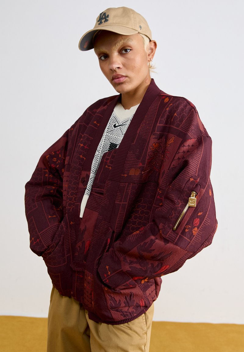 Burgunderfarbene Kimono-Jacke mit aufwendigen floralen und geometrischen Mustern. Sie verfügt über weite Ärmel, Taschen auf der Vorderseite und eine mit Reißverschluss verschließbare Armtasche.