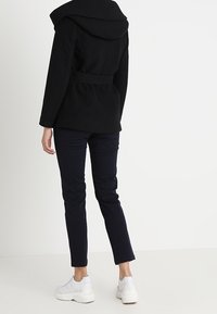 Schwarzer Wollmischmantel mit breitem Kragen und Taillengürtel, kombiniert mit marineblauen Hosen und weißen Sneakern, von hinten betrachtet.