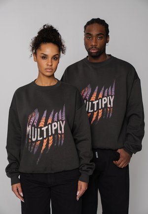 Zwei Personen tragen schwarze Sweatshirts mit bunten Kratzspuren und dem Wort "MULTIPLY" auf der Vorderseite, sie stehen vor einem schlichten Hintergrund.