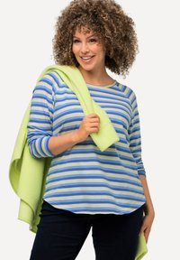 Gestreiftes langärmliges Hemd in Blau und Grün mit abgerundetem Saum. In eine leuchtend grüne Decke gehüllt. Dunkle Jeans vervollständigen das Outfit.