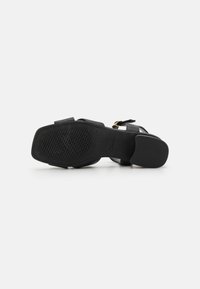 Sandal cu toc negru, cu multiple curele, talpă din cauciuc texturat și un accent din cataramă aurie. Partea superioară din piele netedă demonstrează un design minimalist.