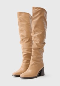 Bottes en cuir marron clair jusqu'au genou avec des talons blocs et un design légèrement affaissé, posées sur un fond blanc uni.