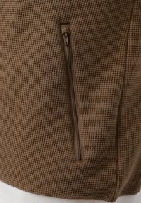 Tissu texturé marron avec une poche zippée verticale, montrant un gros plan détaillé du matériau du vêtement et des coutures.