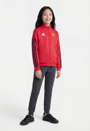 Ung person står og smiler iført en rød Manchester United træningsjakke, mørke joggebukser og sorte sneakers på en hvid baggrund.