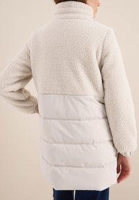 Veste en polaire crème avec un col haut, une partie supérieure texturée et une partie inférieure lisse et matelassée présentant des coutures horizontales.