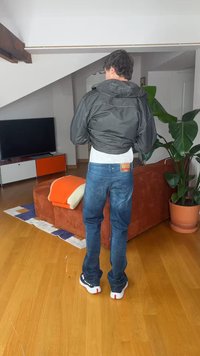 Mann in schwarzer Jacke, blauer Jeans und Sneakers steht mit dem Rücken zur Kamera in einem Wohnzimmer mit Holzboden, orangefarbenem Sofa, Fernseher und großem Topfpflanze.