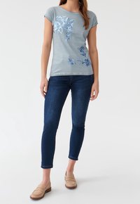Jasnoniebieska koszulka z krótkim rękawem z niebieskim kwiatowym wzorem z cekinów na przodzie. Połączona z ciemnoniebieskimi dżinsami i beżowymi mokasynami.