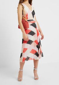 Mujer vistiendo un vestido midi sin mangas y con pliegues, con patrones geométricos en rojo, negro, blanco y rosa, tacones plateados con tiras y un bolso texturizado rojo.