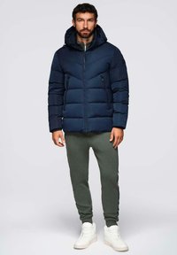Giacca puffer blu navy con zip, cappuccio e due tasche laterali, indossata sopra pantaloni joggers verde oliva e sneakers bianche. Texture liscia.