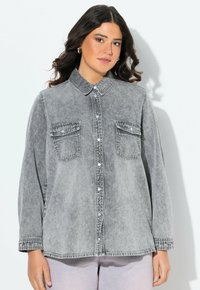 Camicia in denim grigio con colletto classico, chiusura a bottoni, due tasche sul petto e maniche lunghe. Indossata sopra pantaloni chiari.
